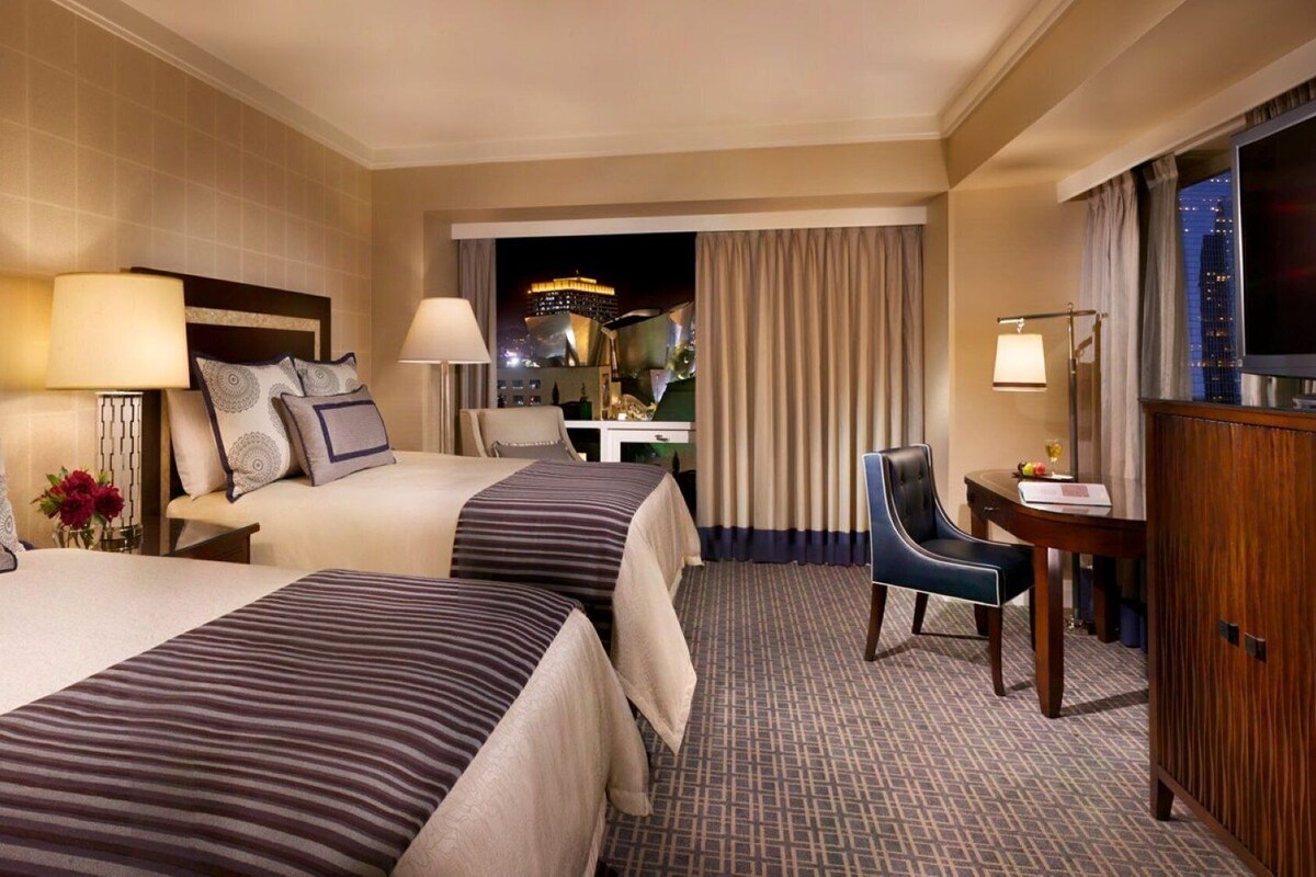 A hotel room with two beds, a desk, a chair, and a TV.