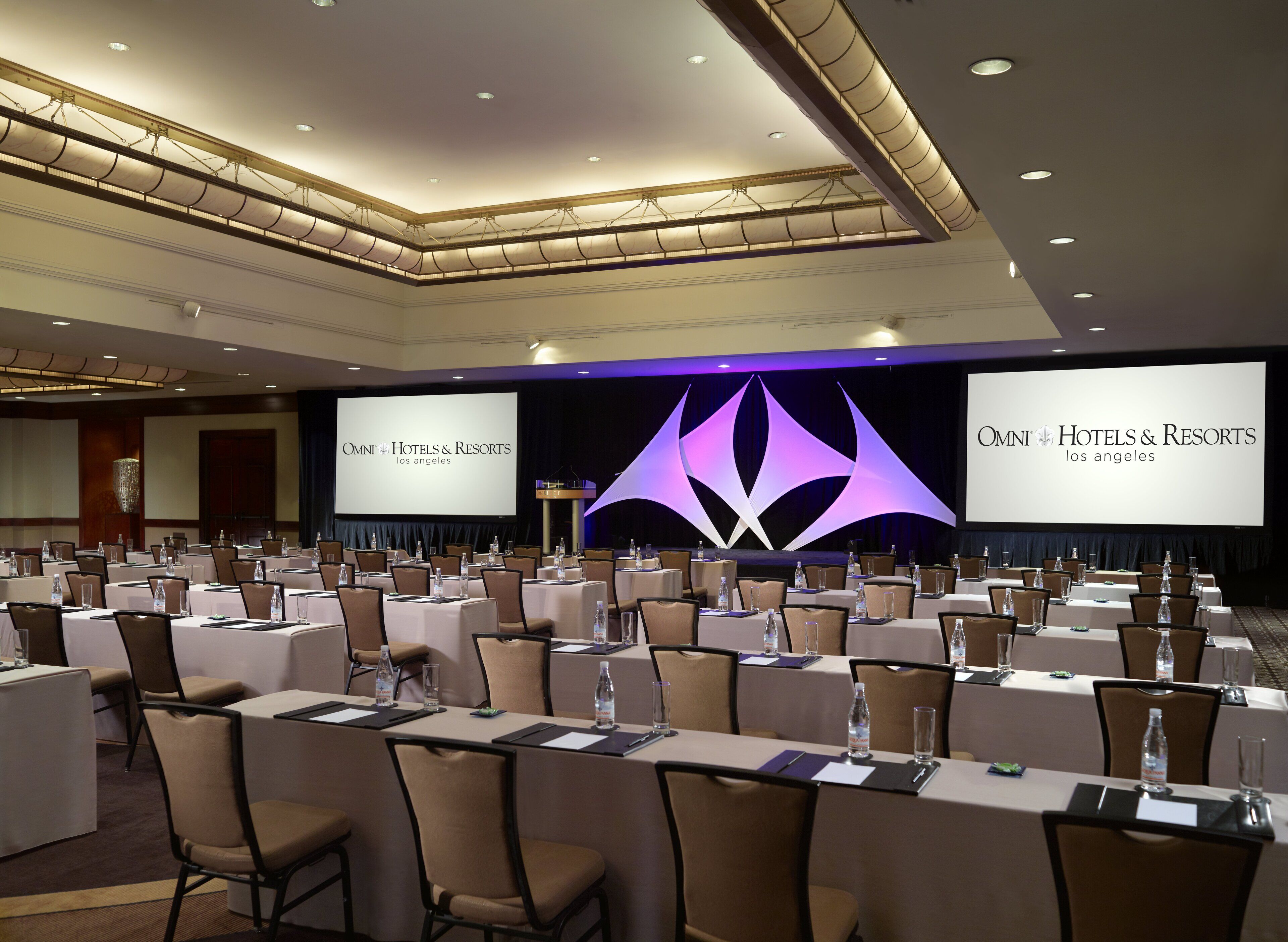 Uma sala de conferências com várias mesas e cadeiras, uma tela grande exibindo o nome do hotel e um palco com um púlpito.