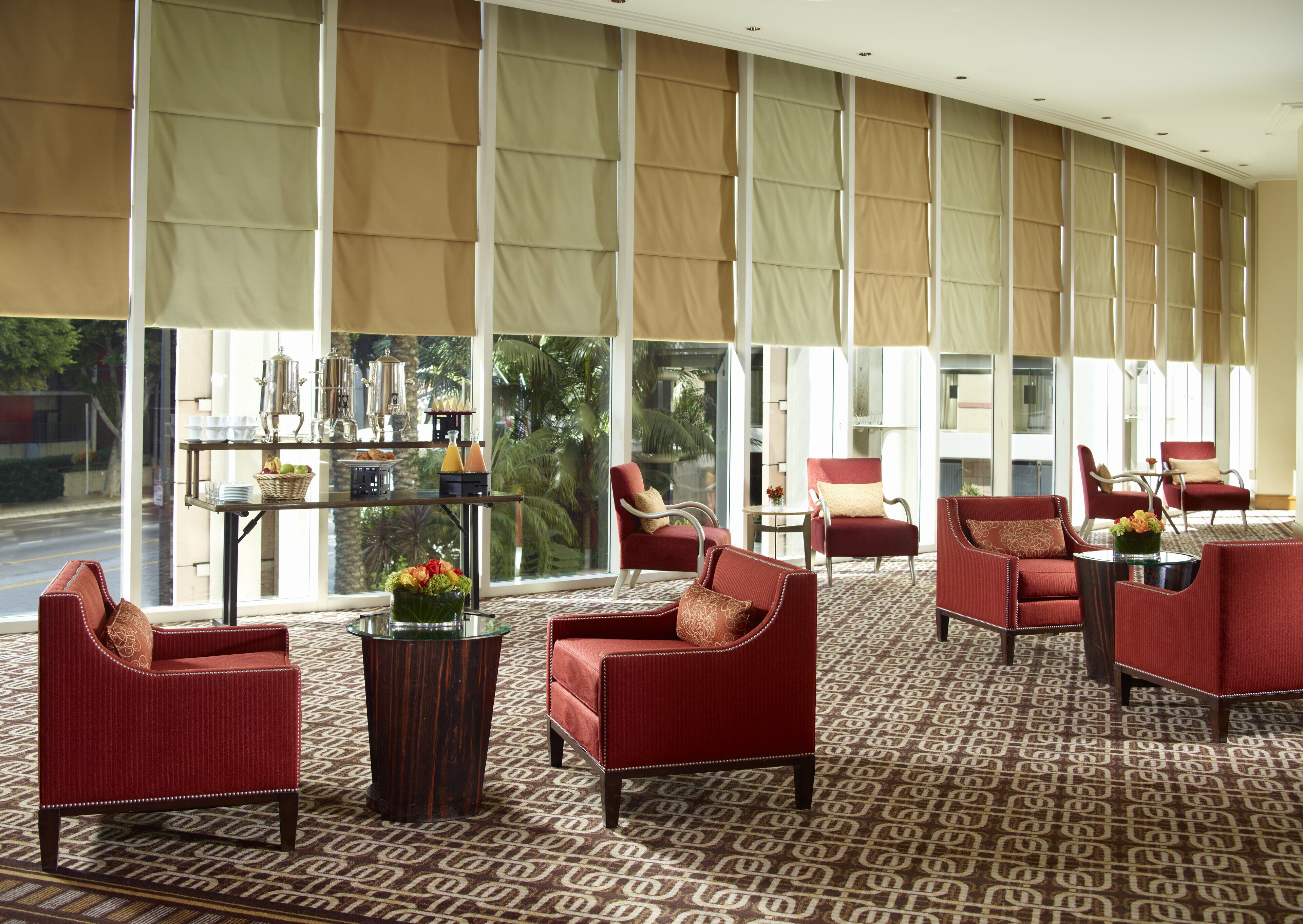 A hotel lobby with red upholstered chairs, a glass coffee table, and a large window with beige curtains.