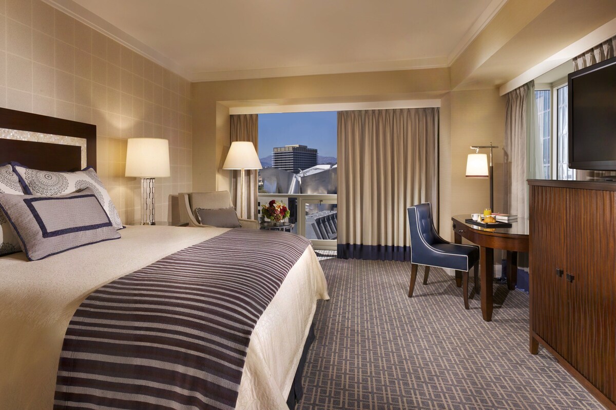 A hotel room with a large bed, two bedside tables with lamps, a desk with a chair, a television, and a view of a building through the window.
