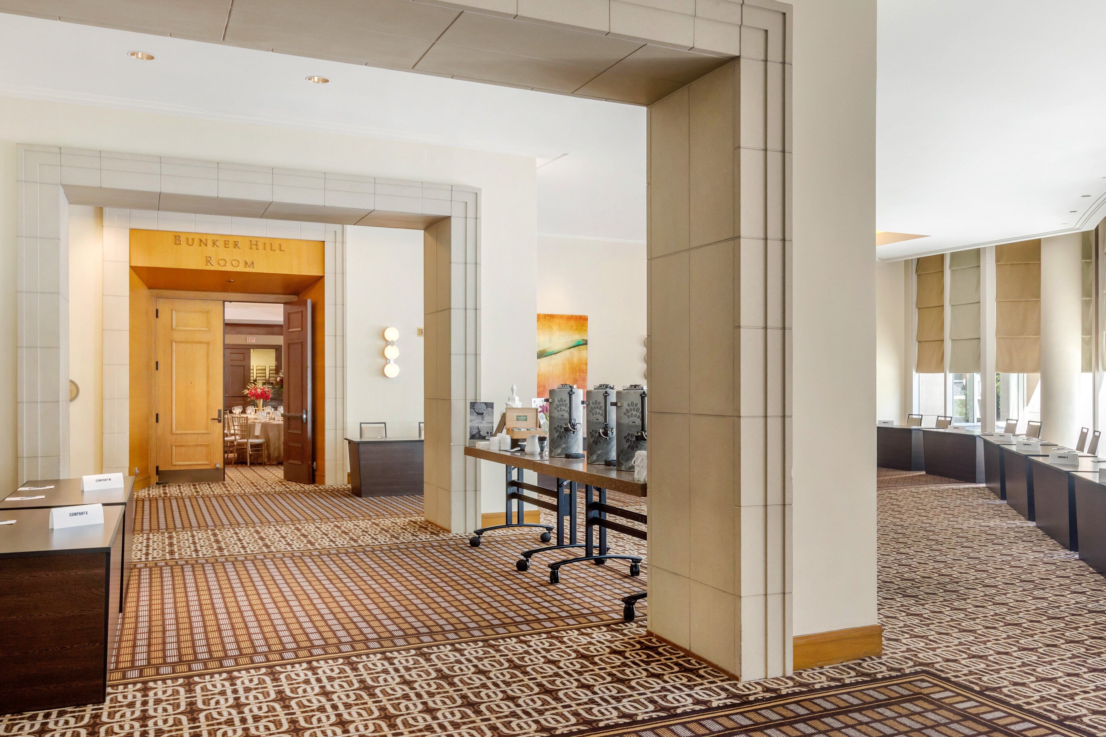 A hotel lobby with a patterned carpet, a reception desk, a Bunker Hill Room sign, and a long table with chairs.