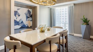 Un comedor moderno con una amplia mesa rectangular, seis sillas, una lámpara decorativa, un cuadro abstracto grande, una planta en maceta y vistas de la ciudad a través de grandes ventanales.