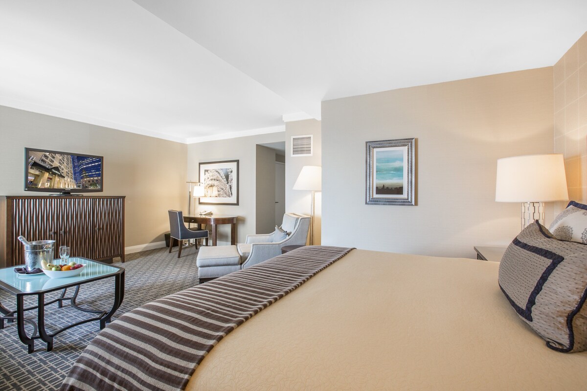 A hotel room with a large bed, a desk with a chair, a small table with a bowl of fruit, and a television.