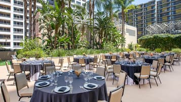 Un área de comedor al aire libre con mesas redondas dispuestas para una comida, rodeada de sillas, con edificios y vegetación al fondo.