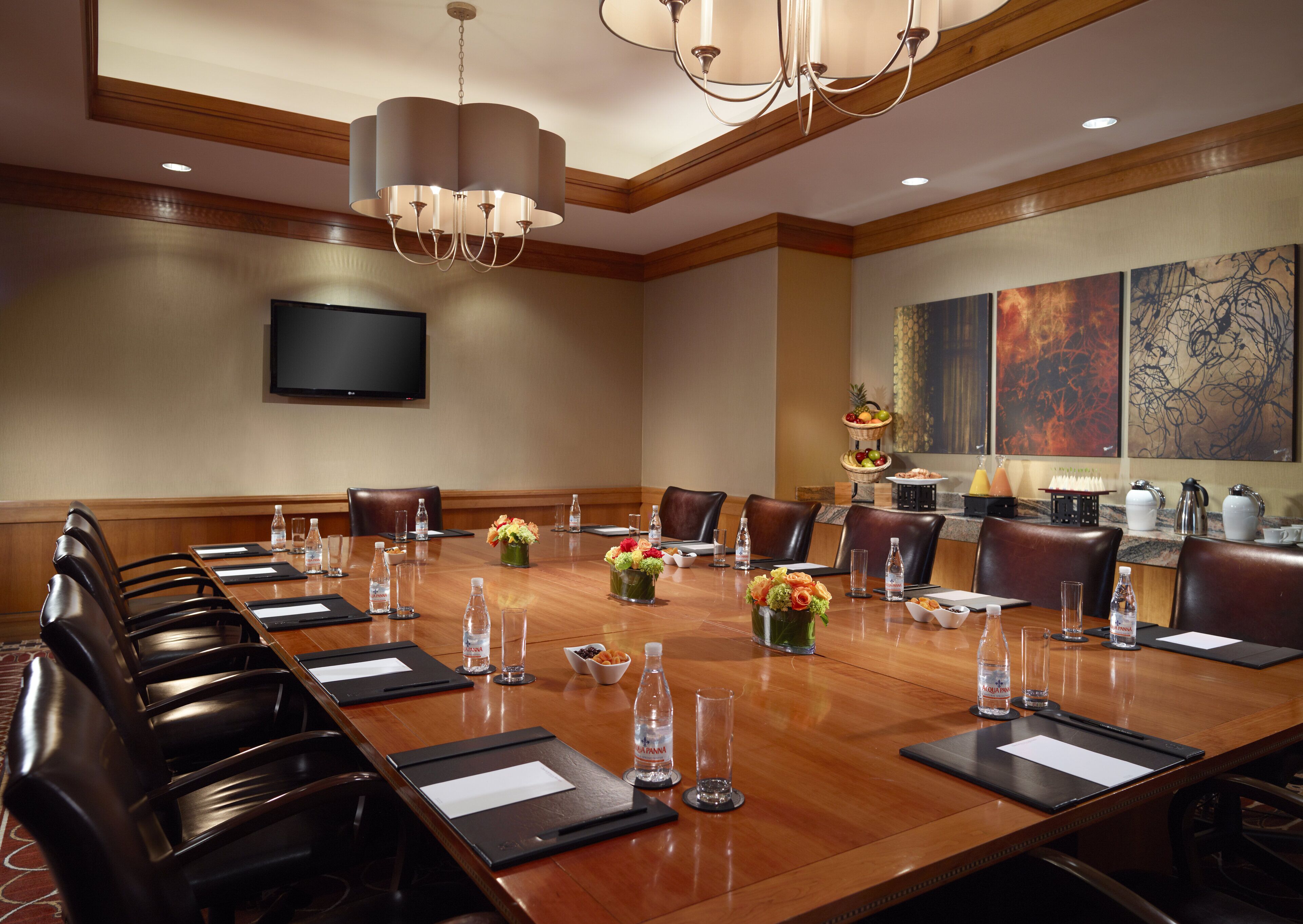 Un salón de conferencias con una larga mesa de madera, sillas de cuero, botellas de agua y centros de mesa con flores.