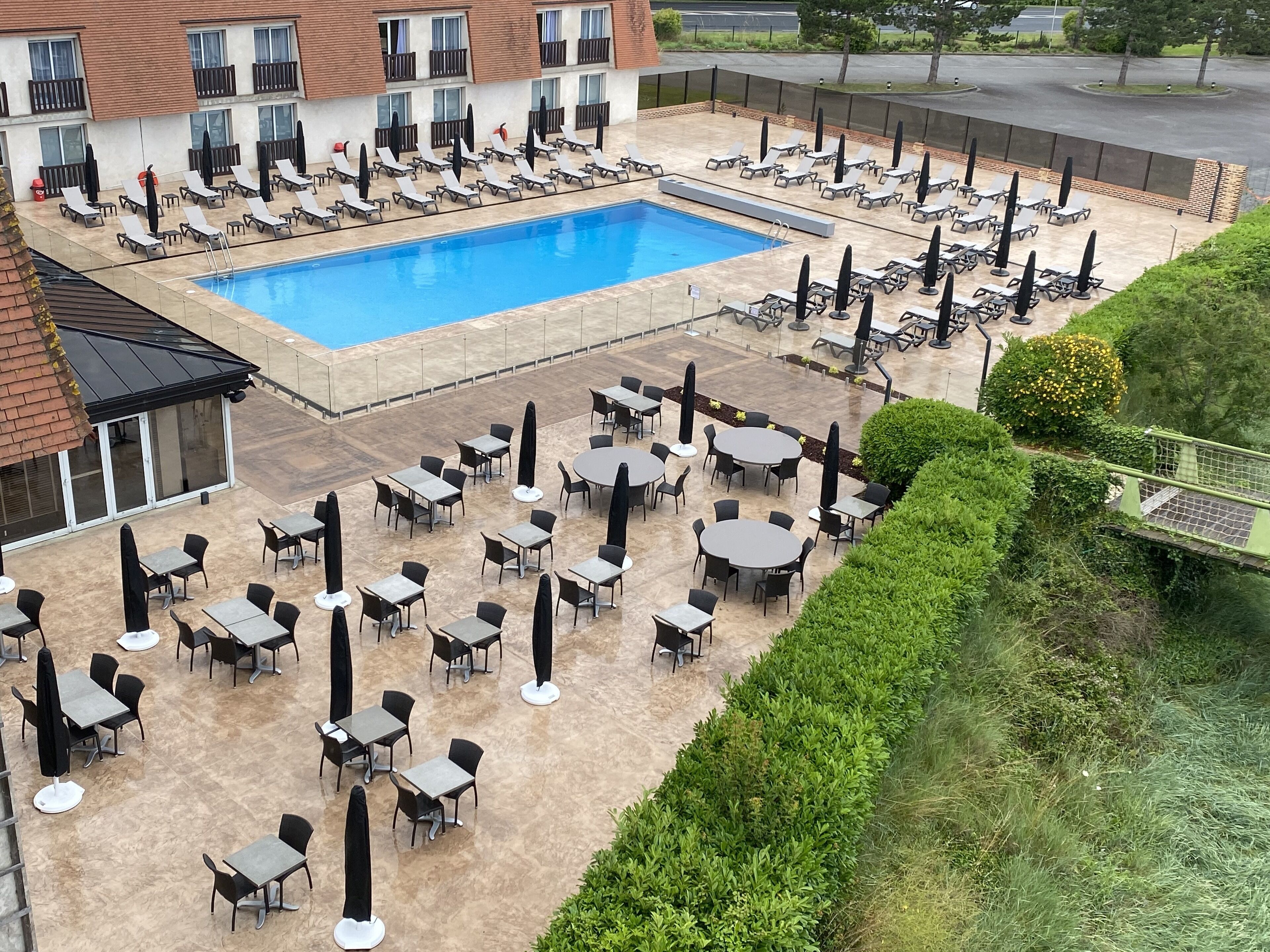 Zona junto a la piscina con tumbonas y mesas, rodeada de follaje y un edificio con balcones.
