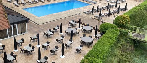 Zona junto a la piscina con tumbonas y mesas, rodeada de follaje y un edificio con balcones.