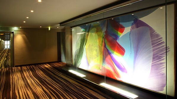 A hallway with a large glass wall displaying a colorful abstract artwork. The floor has a striped carpet pattern.