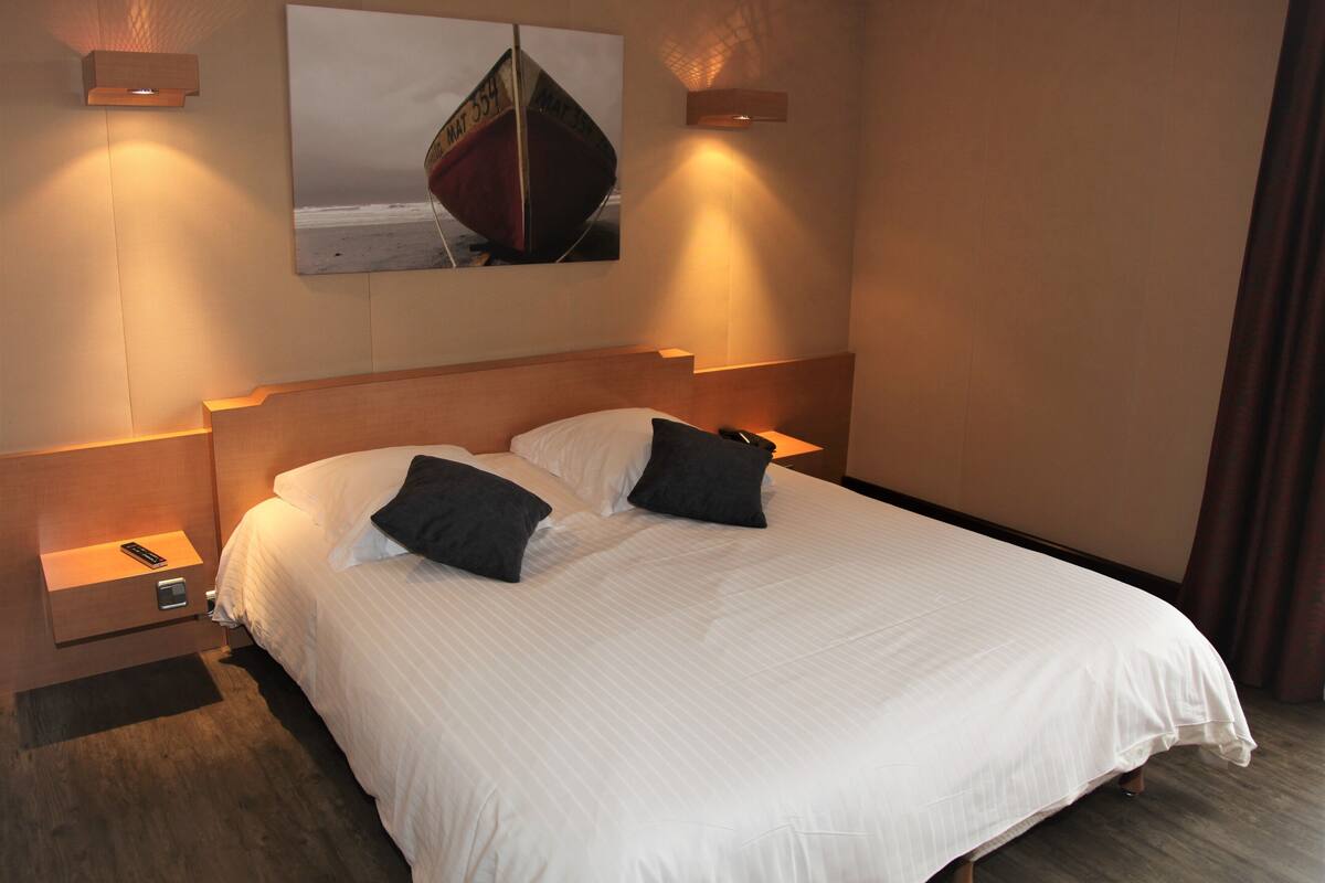 A neatly made bed with white linens and two pillows, a wooden headboard, bedside lamps, and a framed picture of a boat on the wall.
