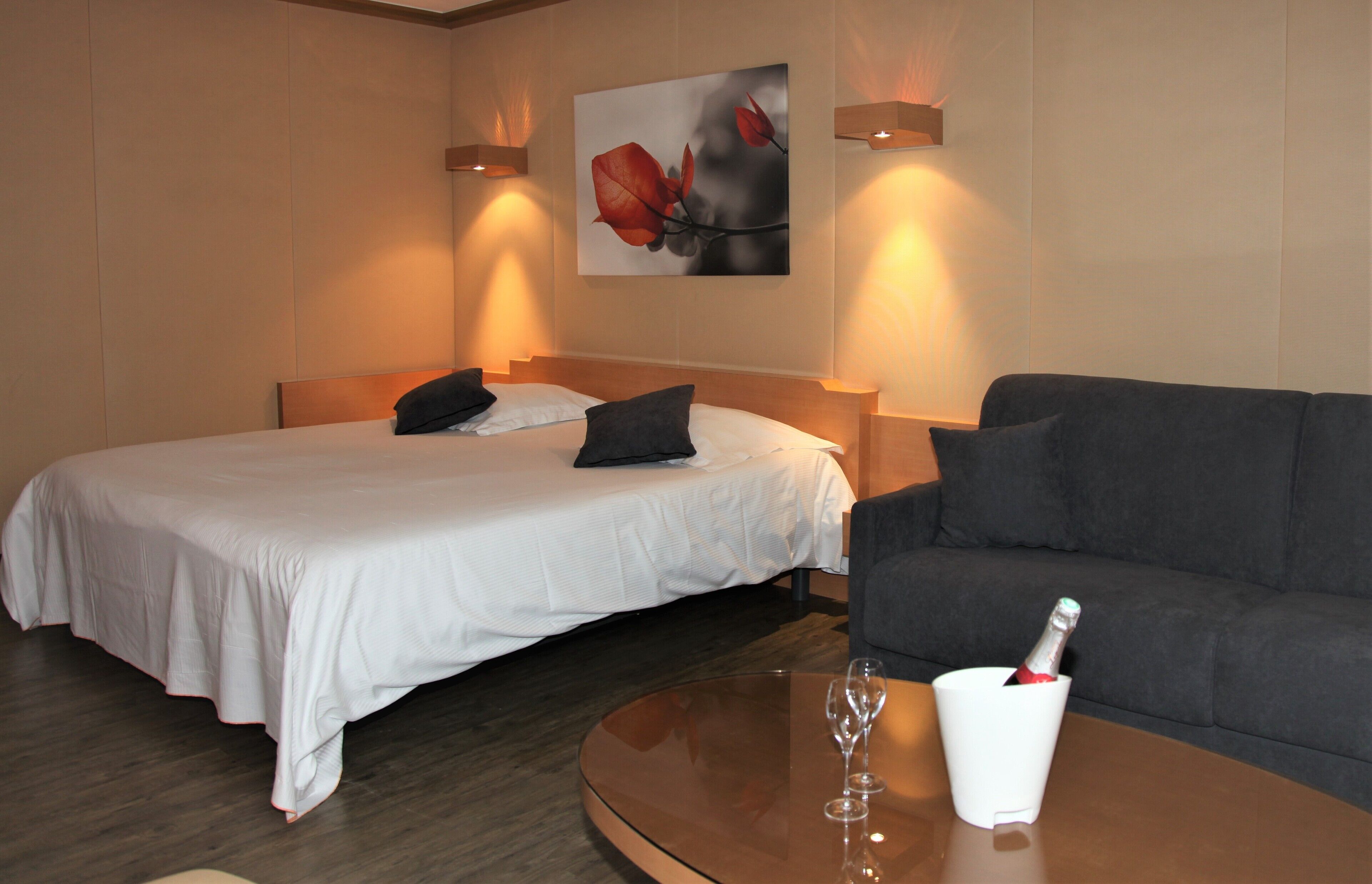 A hotel room with a bed, a sofa, a glass table with a wine glass and a bottle, and a painting on the wall.