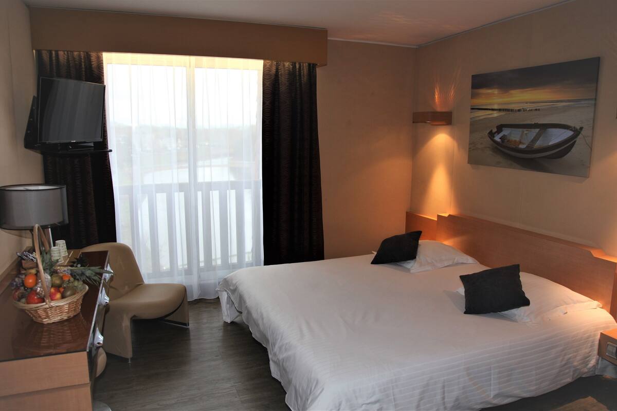 A hotel room with a large bed, a desk with a lamp, a chair, a TV, and a painting of a boat on the wall.