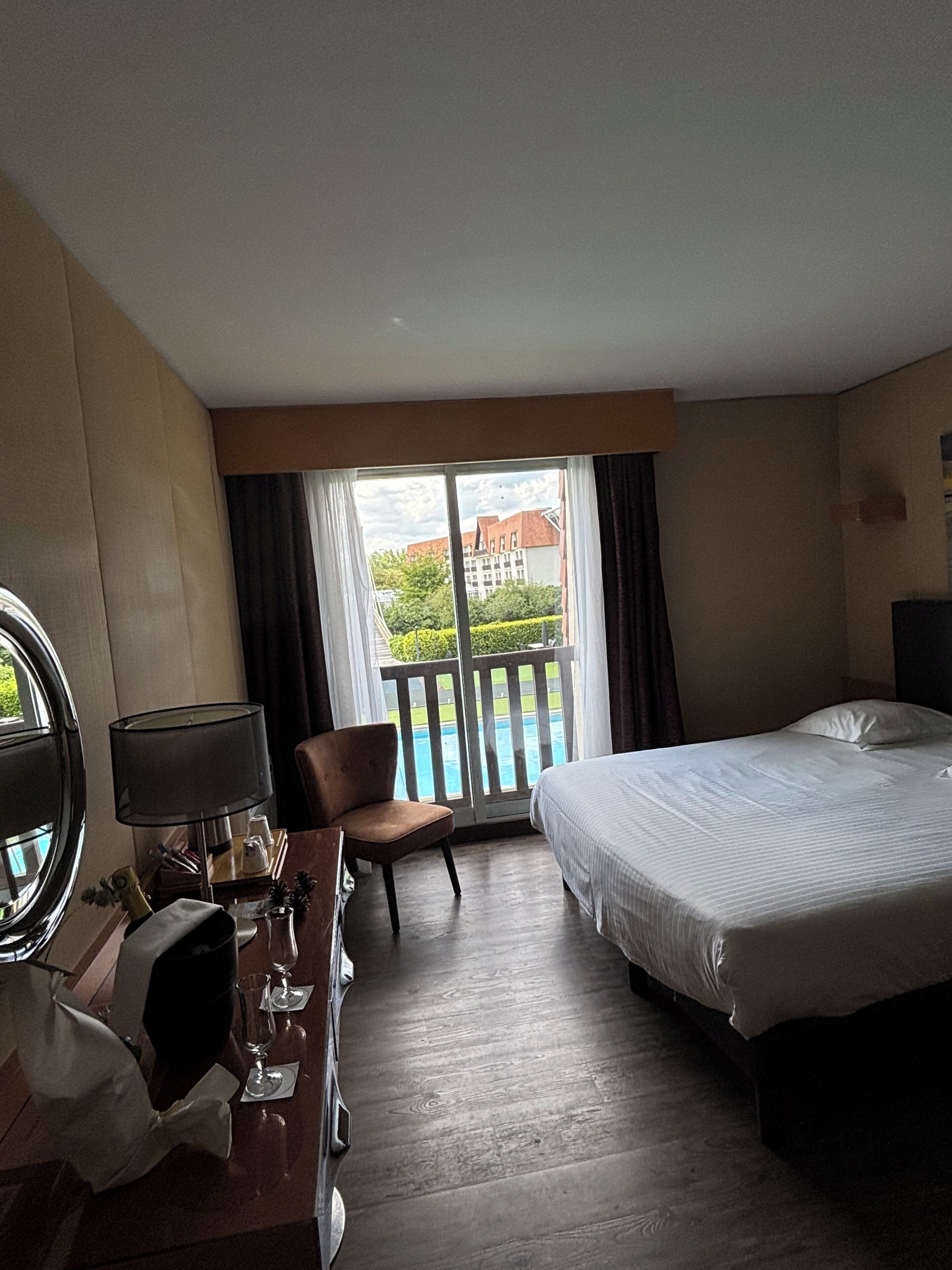 Ein Hotelzimmer mit einem Bett, einem Schreibtisch mit Lampe, einem Sessel und einem Fenster mit Blick auf einen Pool und Gebäude.