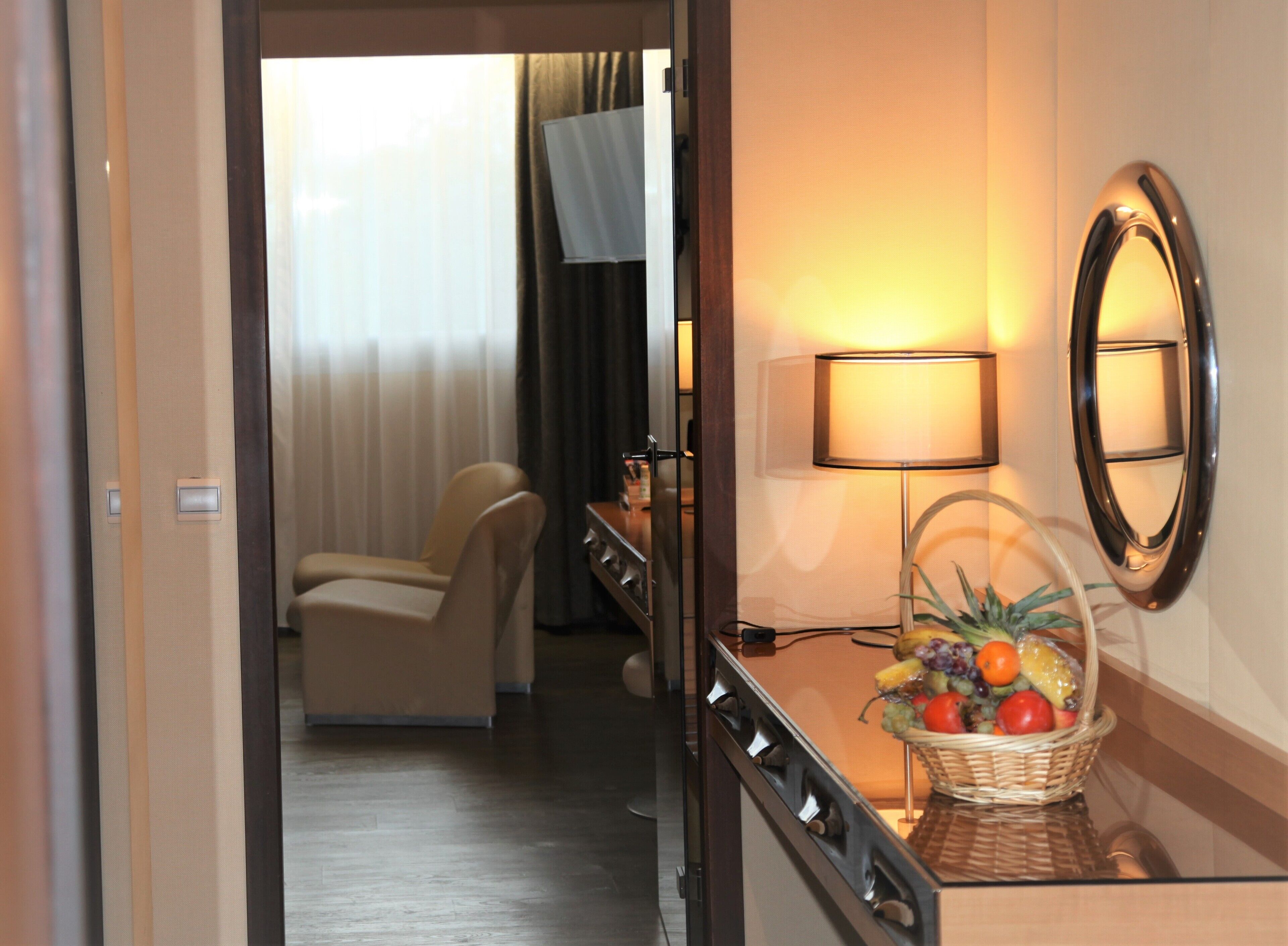 A hotel room with a bed, a desk with a lamp, a fruit basket, and a mirror on the wall.