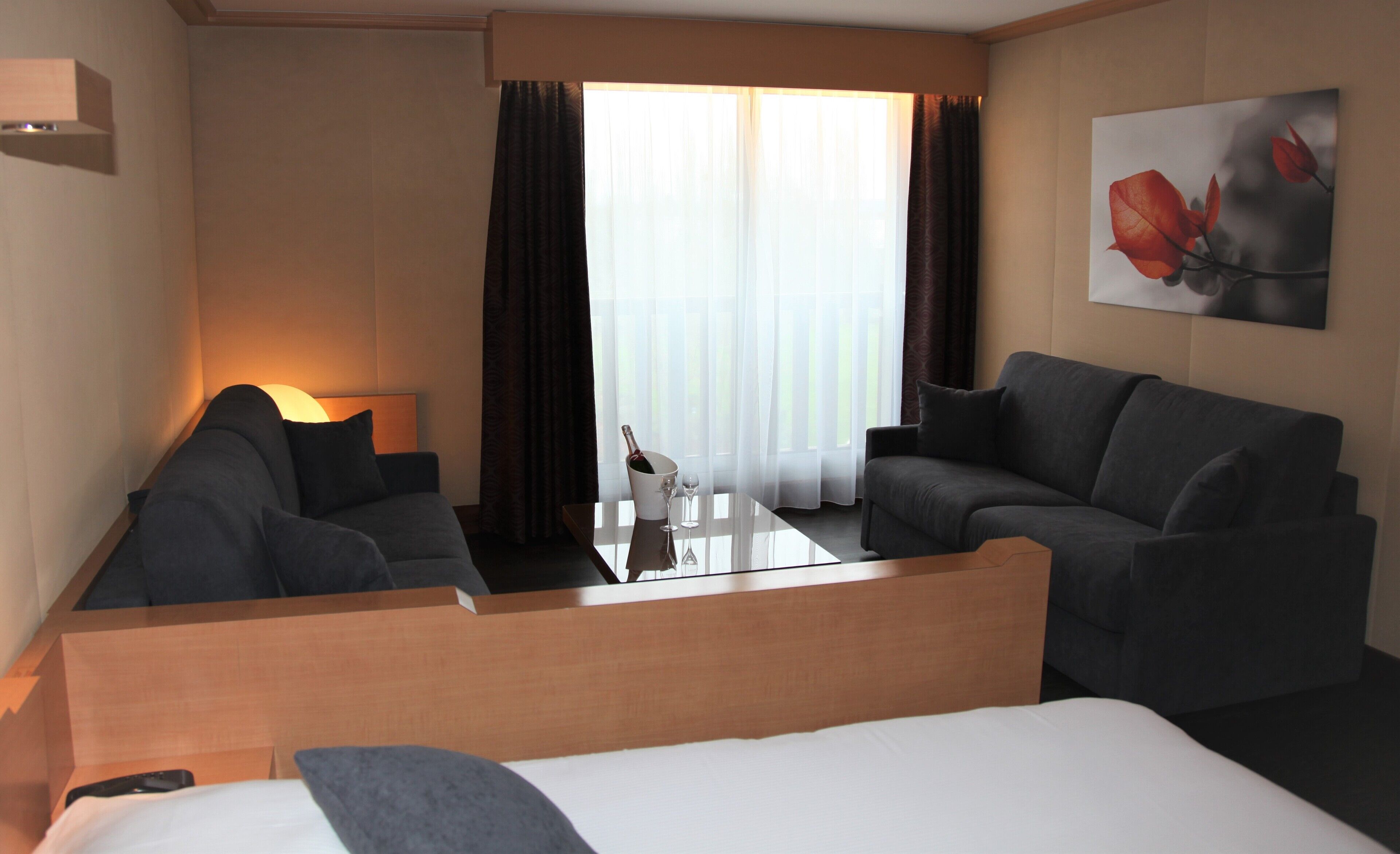 A hotel room with a bed, two armchairs, a small table with a bottle and glasses, a lamp, and a painting on the wall.