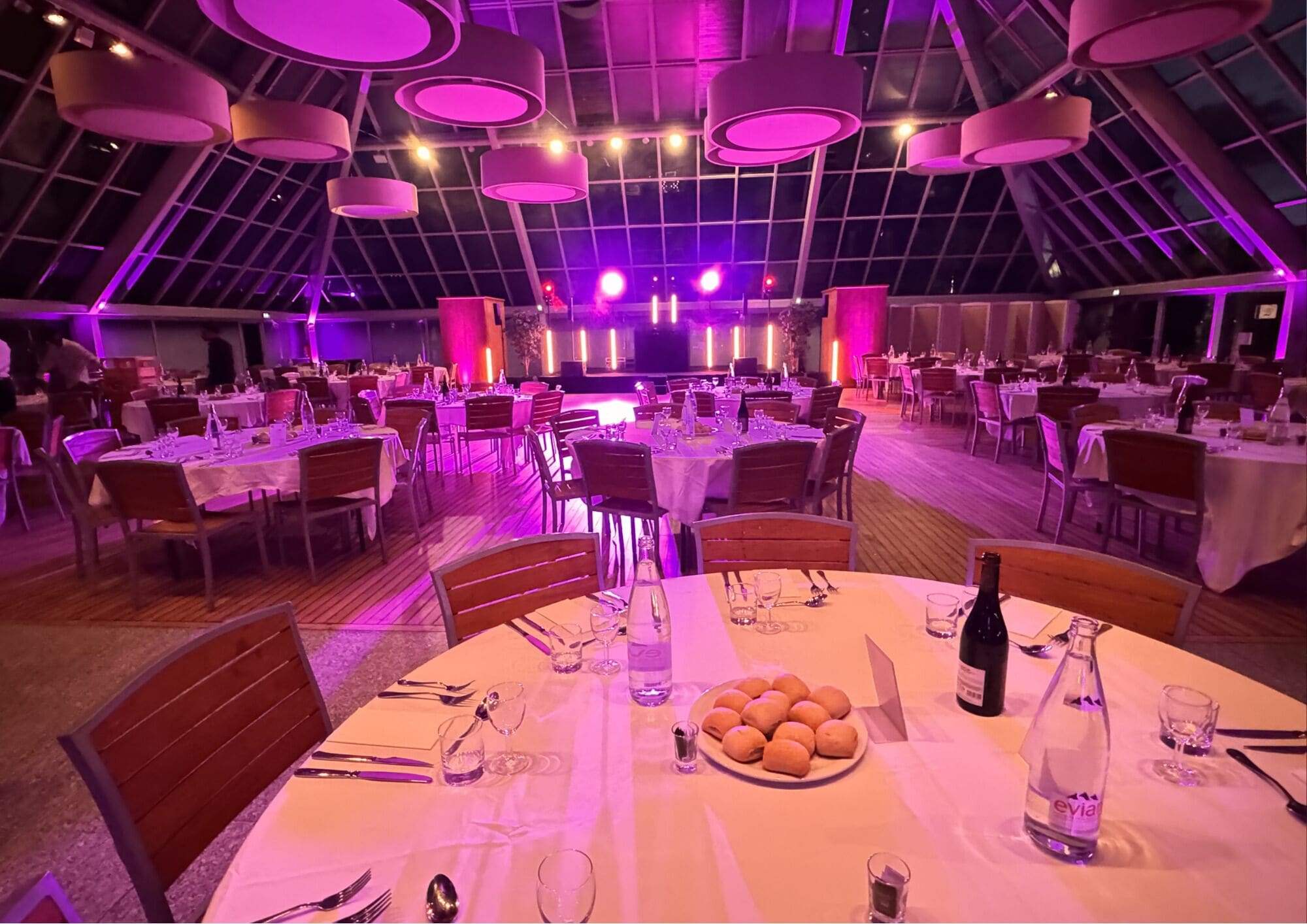 A large indoor dining area with tables set for multiple guests, a stage in the background, and a high ceiling with hanging lights.