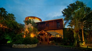 A two-story building with a circular structure on the roof, surrounded by lush greenery and palm trees.