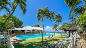 Un espace piscine avec des chaises longues, un coin salon abrité et des palmiers.