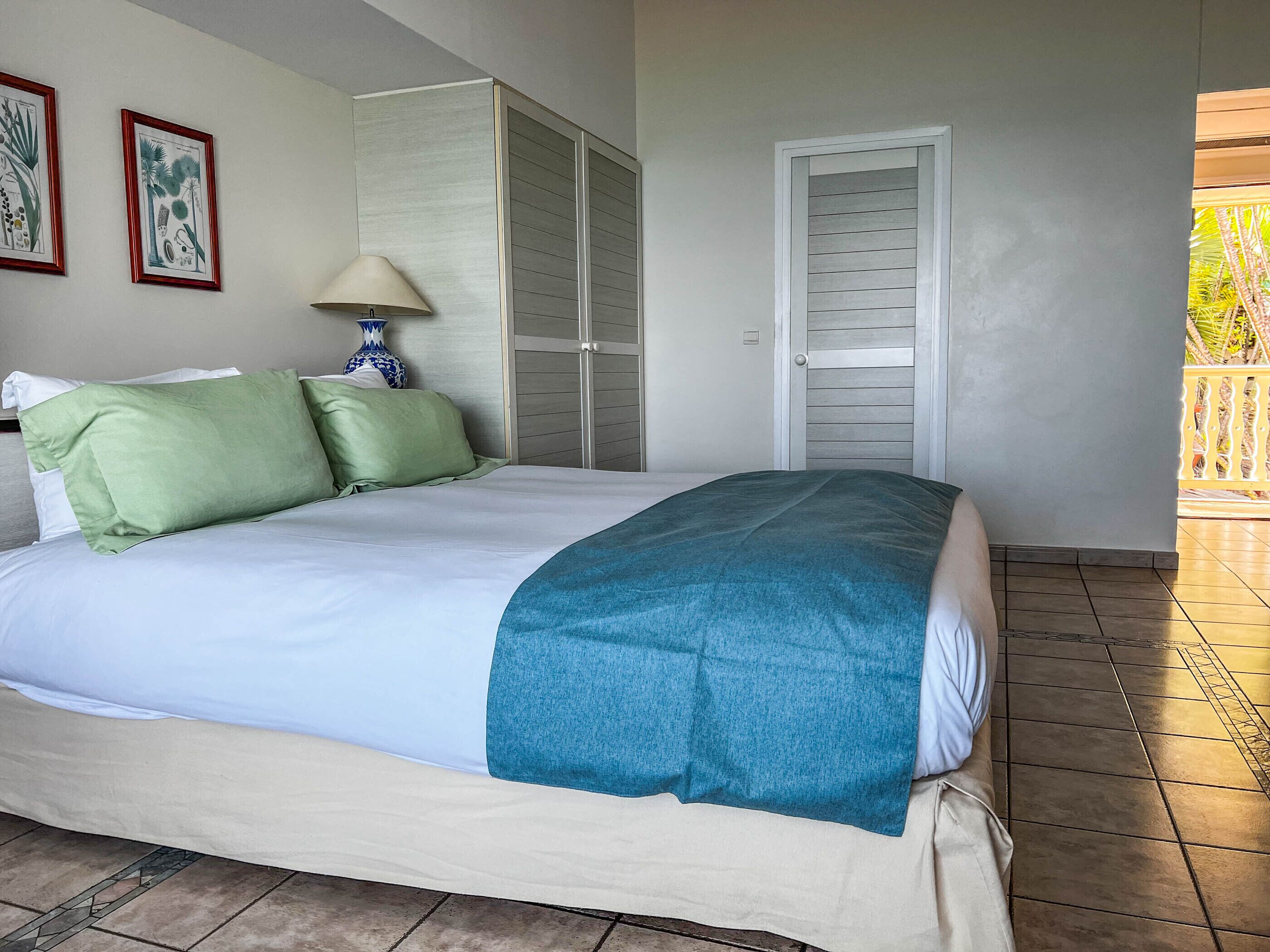 Una cama bien hecha con una manta color turquesa, un almohadón verde y un almohadón blanco. Hay una lámpara con un jarrón azul sobre una mesita de noche. Una puerta corrediza da acceso a un balcón con barandilla.