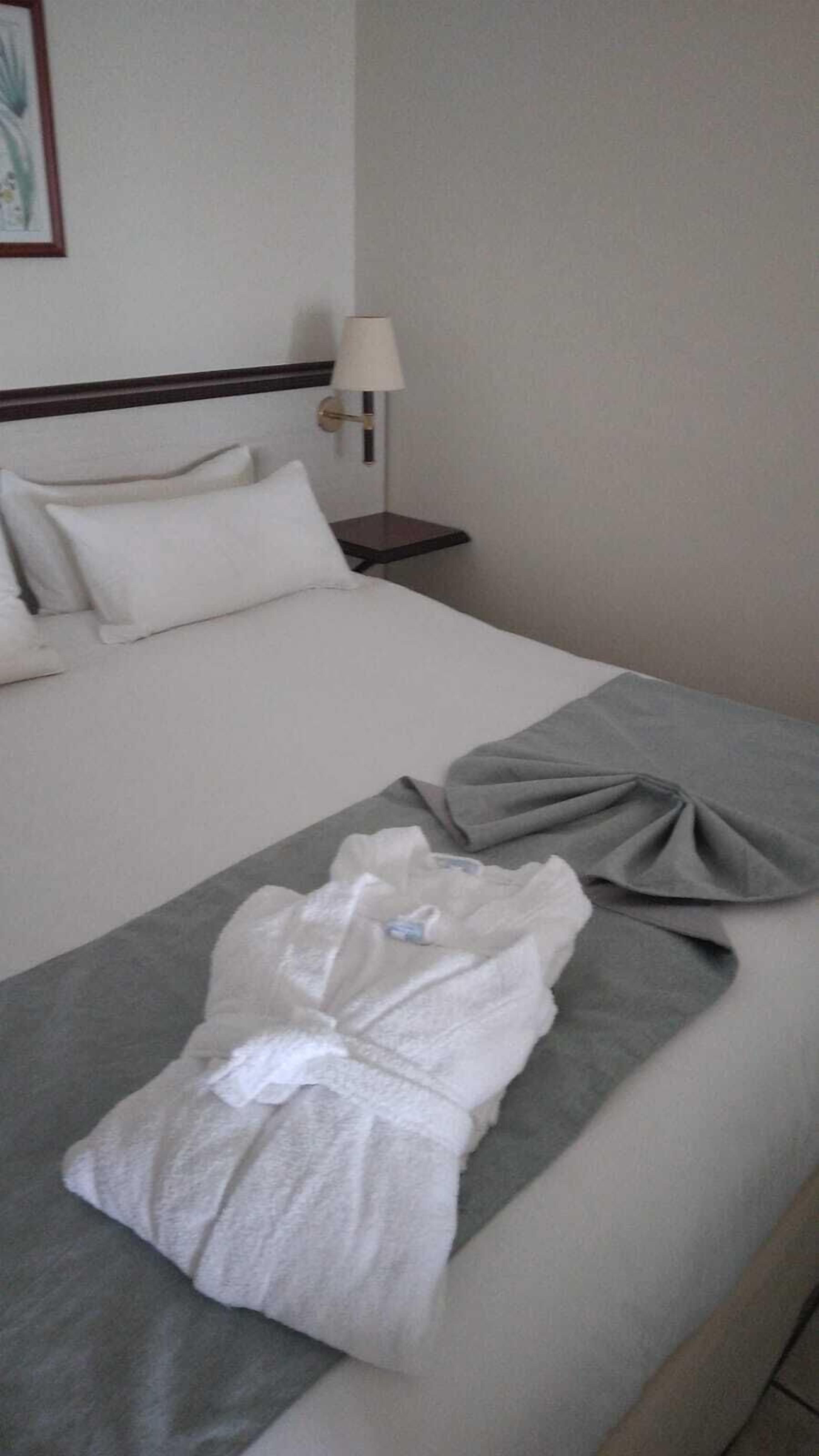 A neatly made bed with white bedding, a grey blanket, and a white robe folded on the bed.