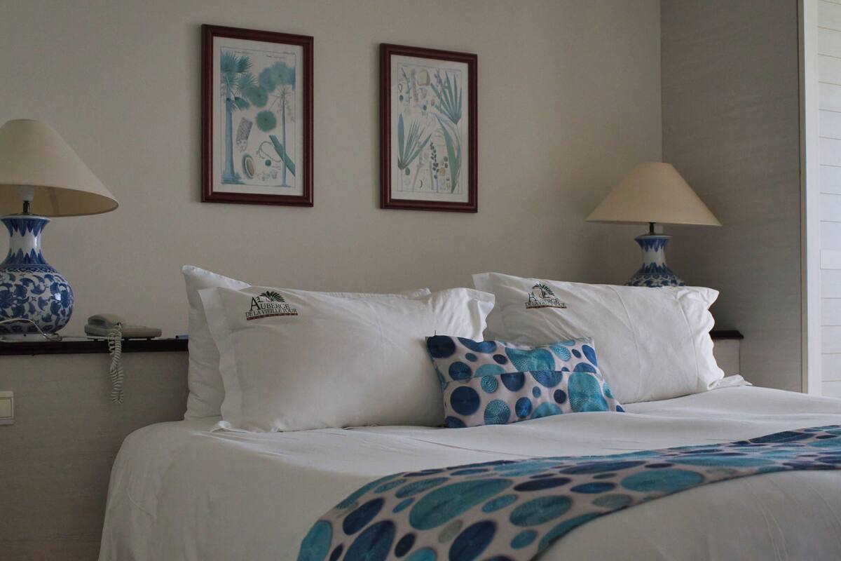 Une chambre d’hôtel comprenant un lit, deux lampes fixées au mur, un téléphone et deux reproductions encadrées de motifs botaniques.