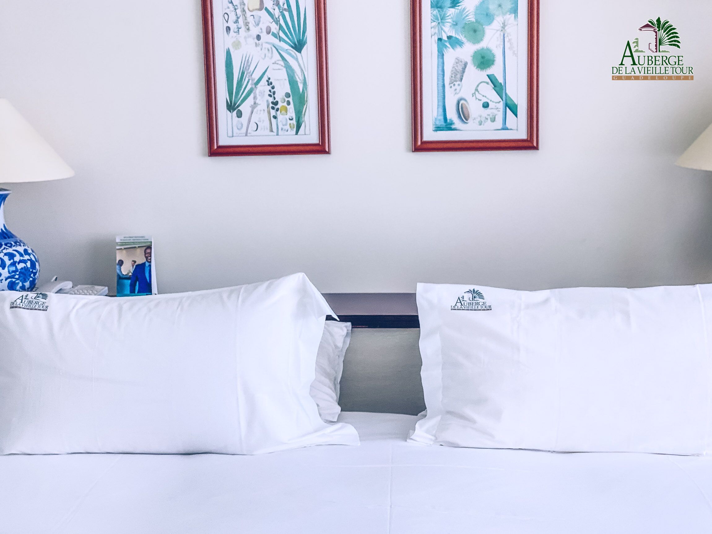 A hotel room with two white beds, a nightstand, a blue vase, and two framed botanical prints on the wall.