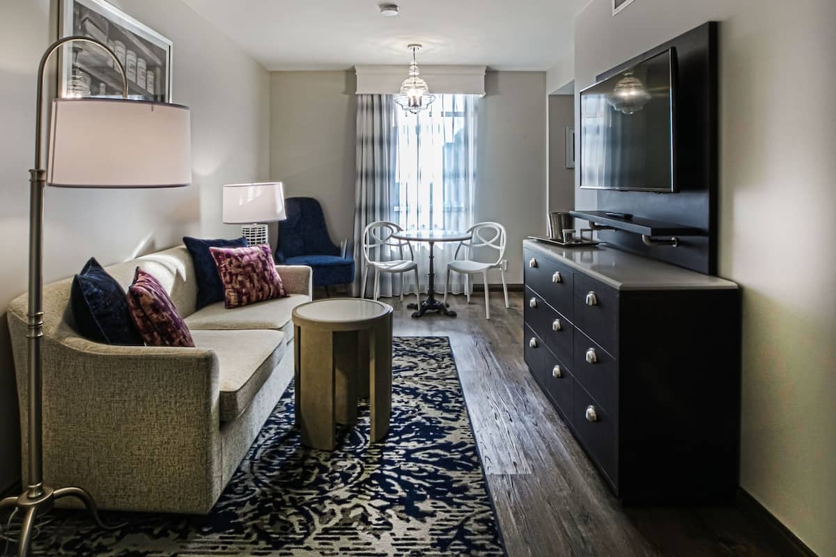 A compact hotel room with a sofa, a small round table, a dresser, a TV, and a lamp.