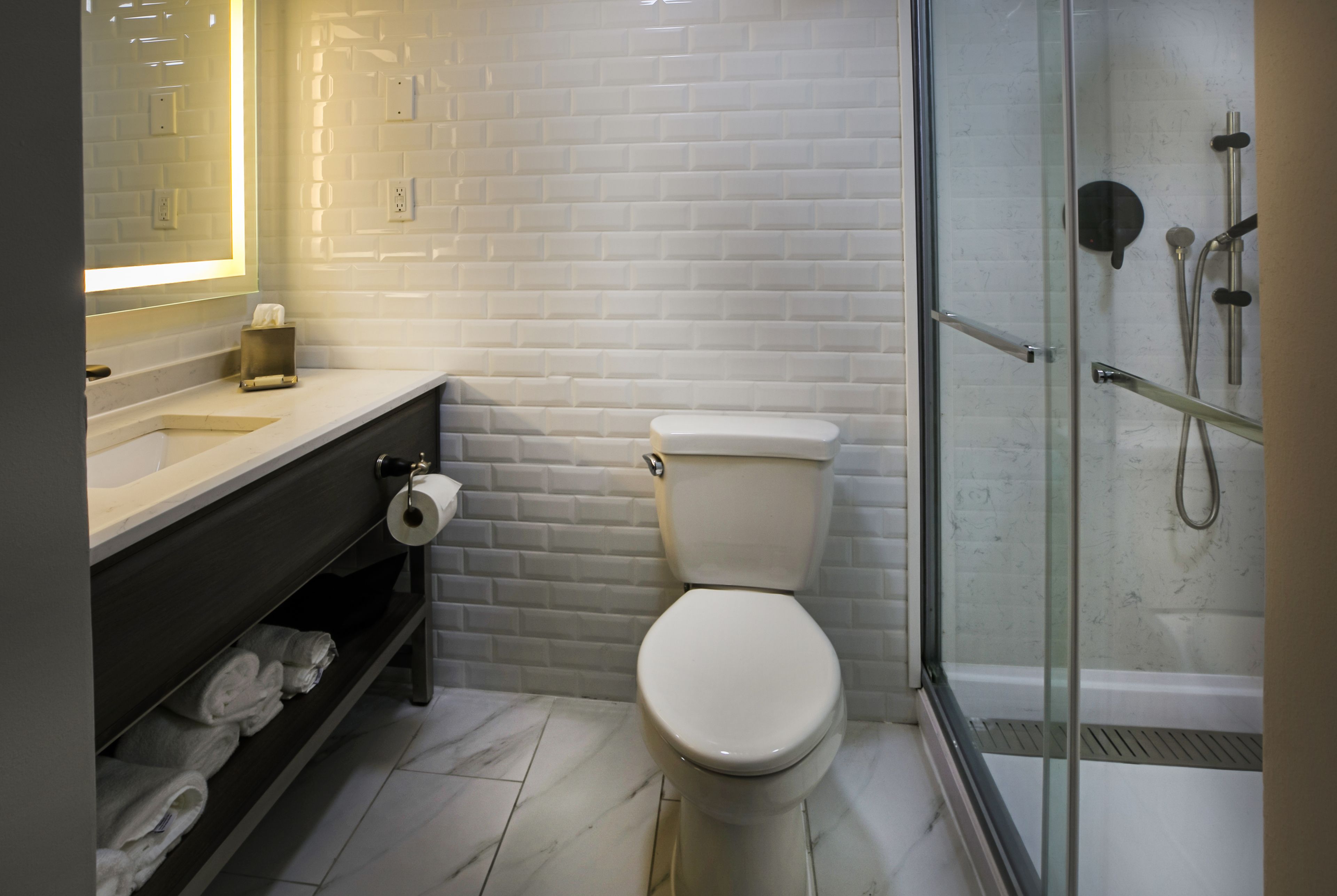 Un baño con inodoro blanco, cabina de ducha con mampara de cristal, pared de azulejos blancos y suelo de mármol.