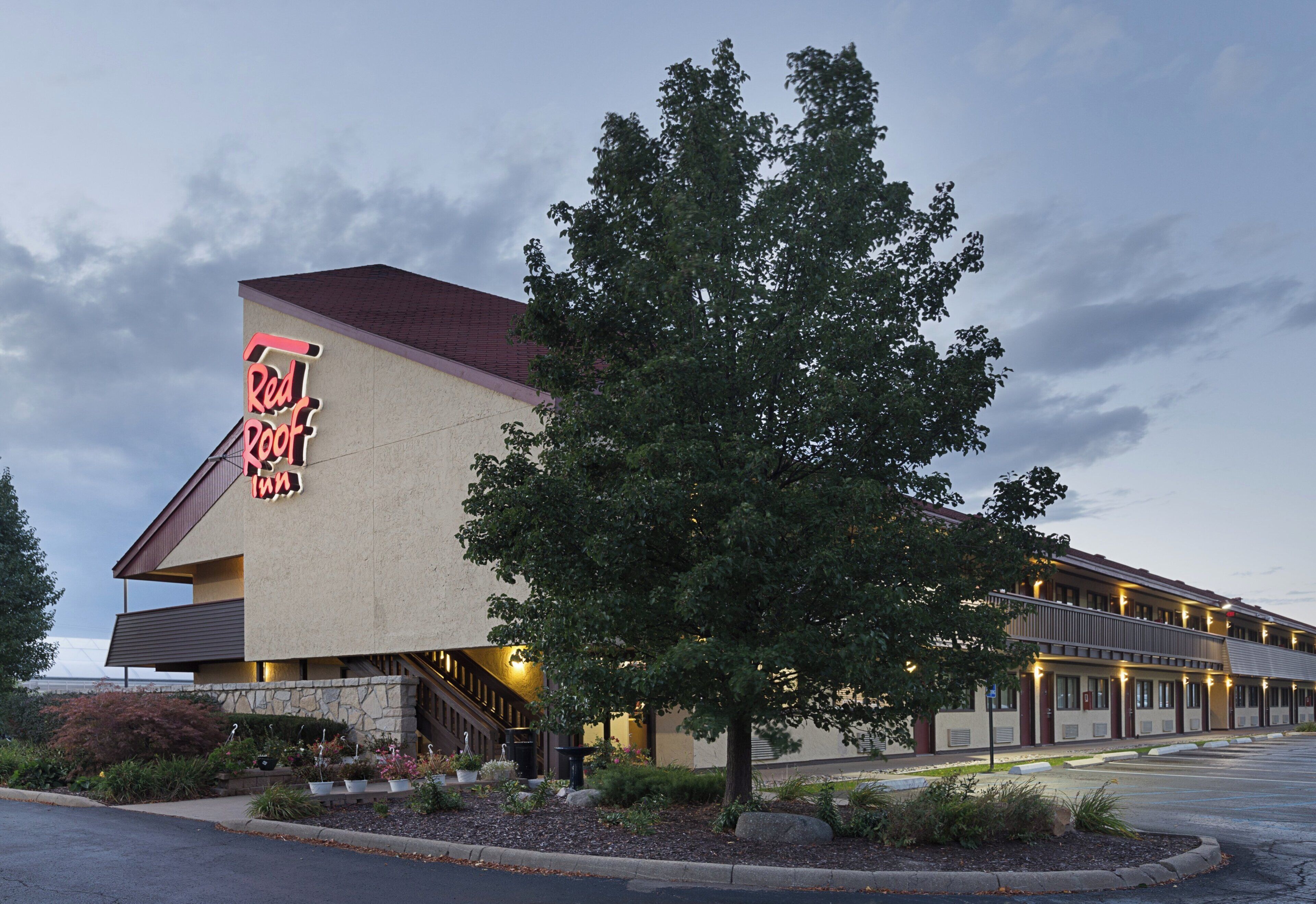 A Red Roof Inn with a large tree in front, a parking lot, and a clear sky.