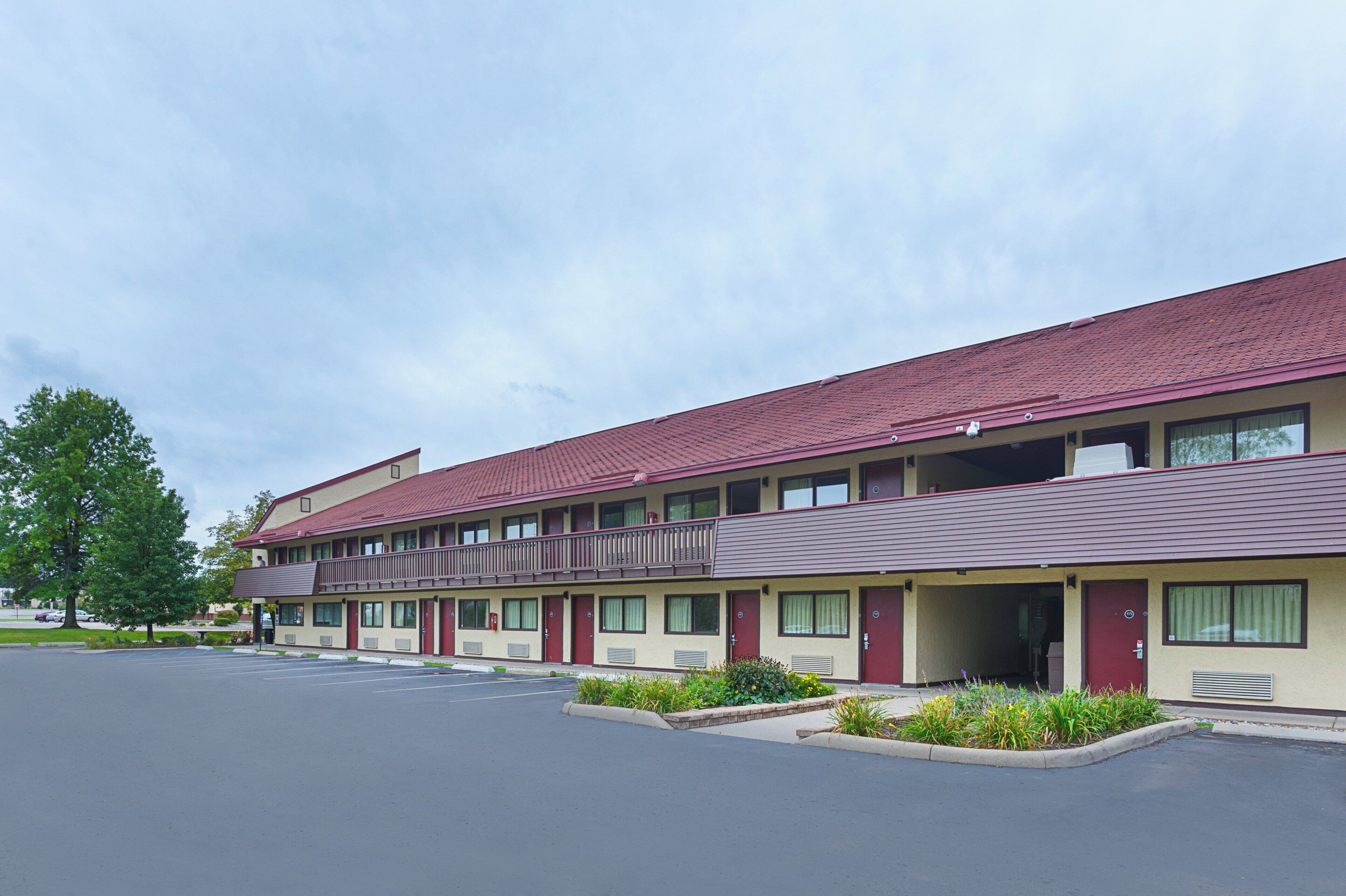 A multi-story motel with a red roof and beige walls, featuring a parking lot and a landscaped area with shrubs.