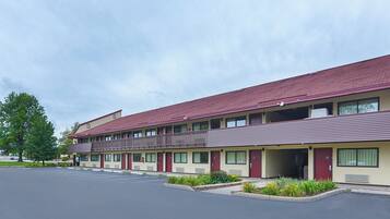 A multi-story motel with a red roof and beige walls, featuring a parking lot and a landscaped area with shrubs.