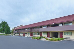 A multi-story motel with a red roof and beige walls, featuring a parking lot and a landscaped area with shrubs. - Red Roof Inn Lansing West - MSU (Lansing)