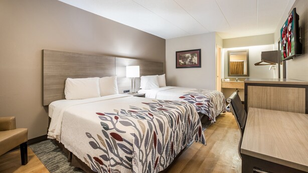 A hotel room with two beds, a desk, a chair, a TV, and a bathroom visible through an open door. - Red Roof Inn Lansing West - MSU (Lansing)