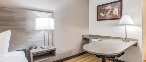 A hotel room with a bed, bedside table, lamp, desk, chair, and a framed picture on the wall.