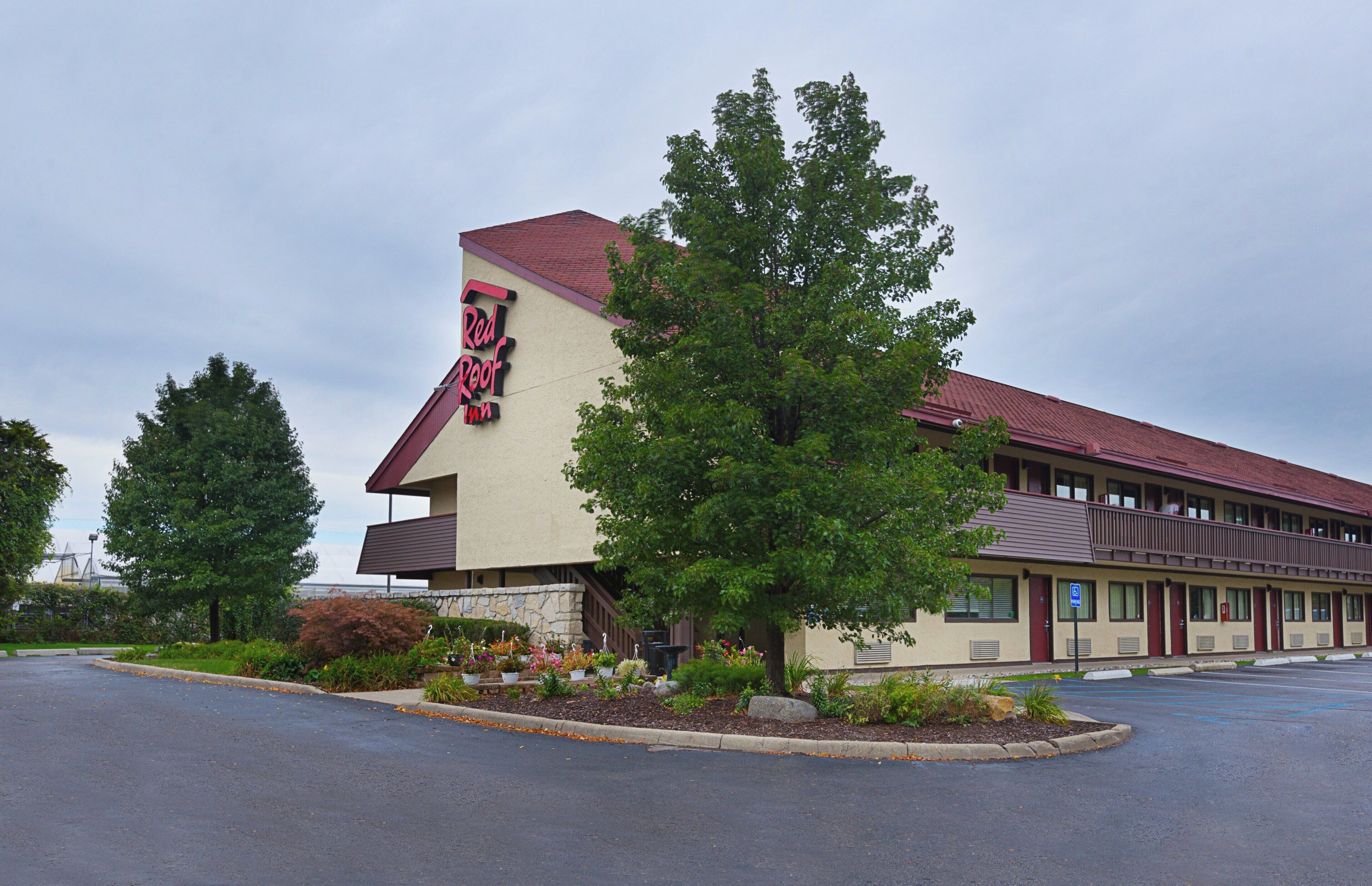 A Red Roof Inn with a red roof, beige exterior, and a parking lot with trees and shrubs.