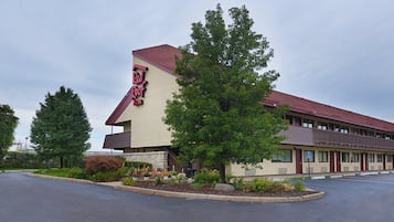 A Red Roof Inn with a red roof, beige exterior, and a parking lot with trees and shrubs.