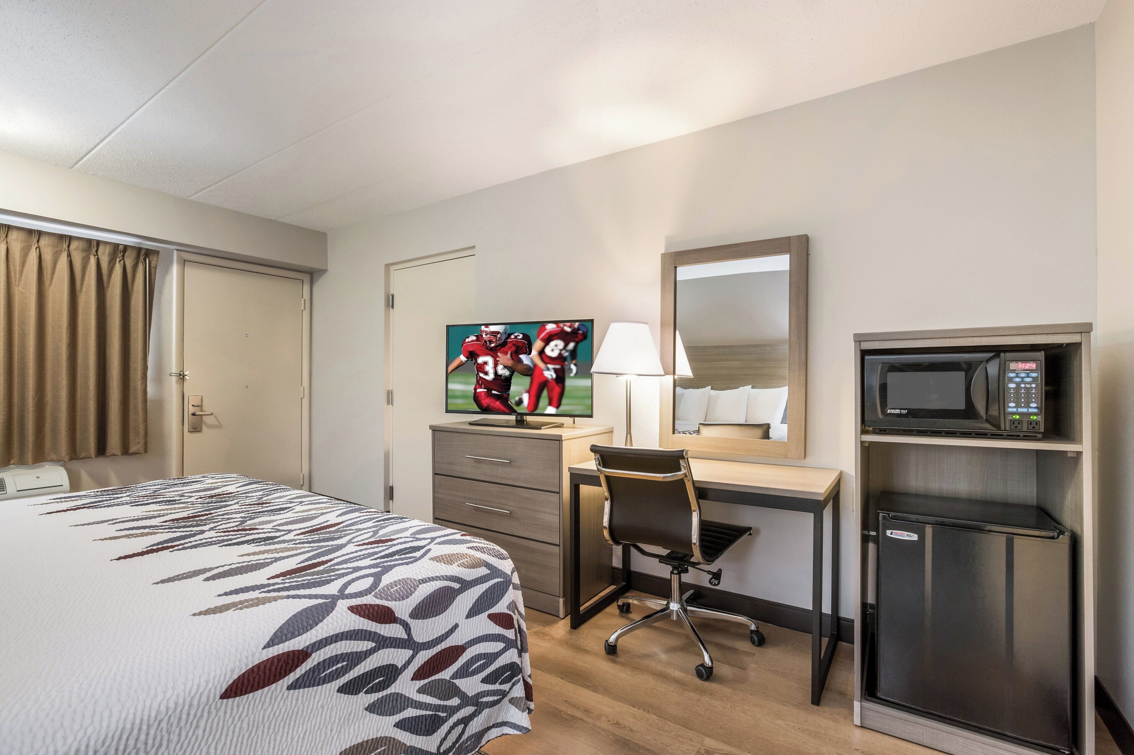 Quarto de hotel com cama, escrivaninha, cadeira, televisão, espelho e minibar.