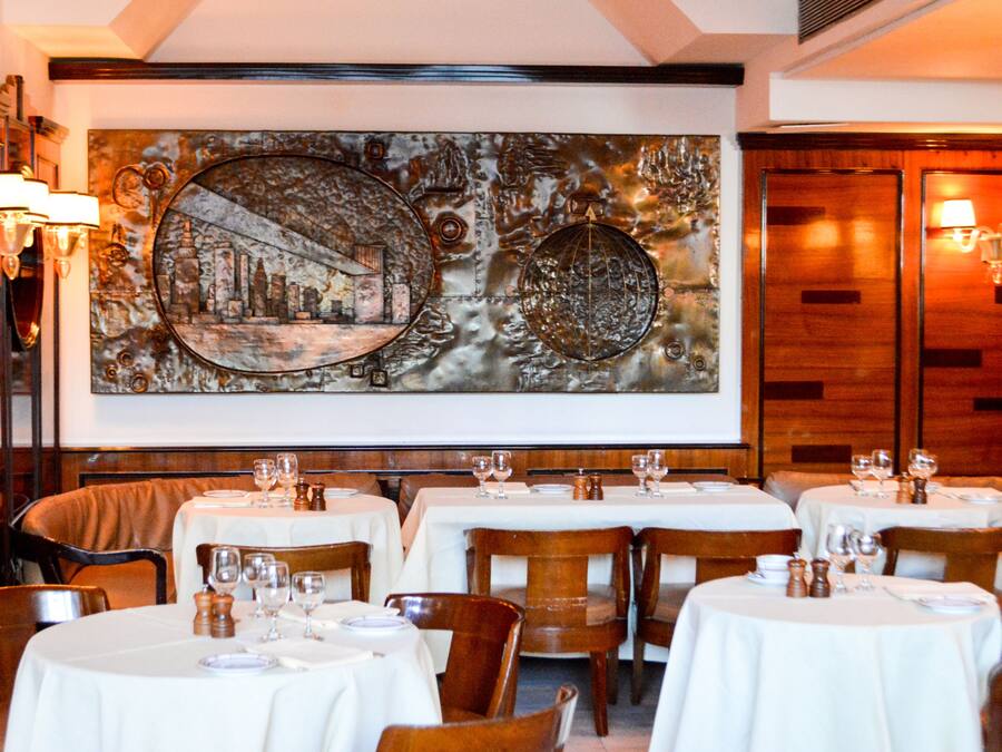 A restaurant with round tables, white tablecloths, and wooden chairs. There is a large wall art piece above the tables.