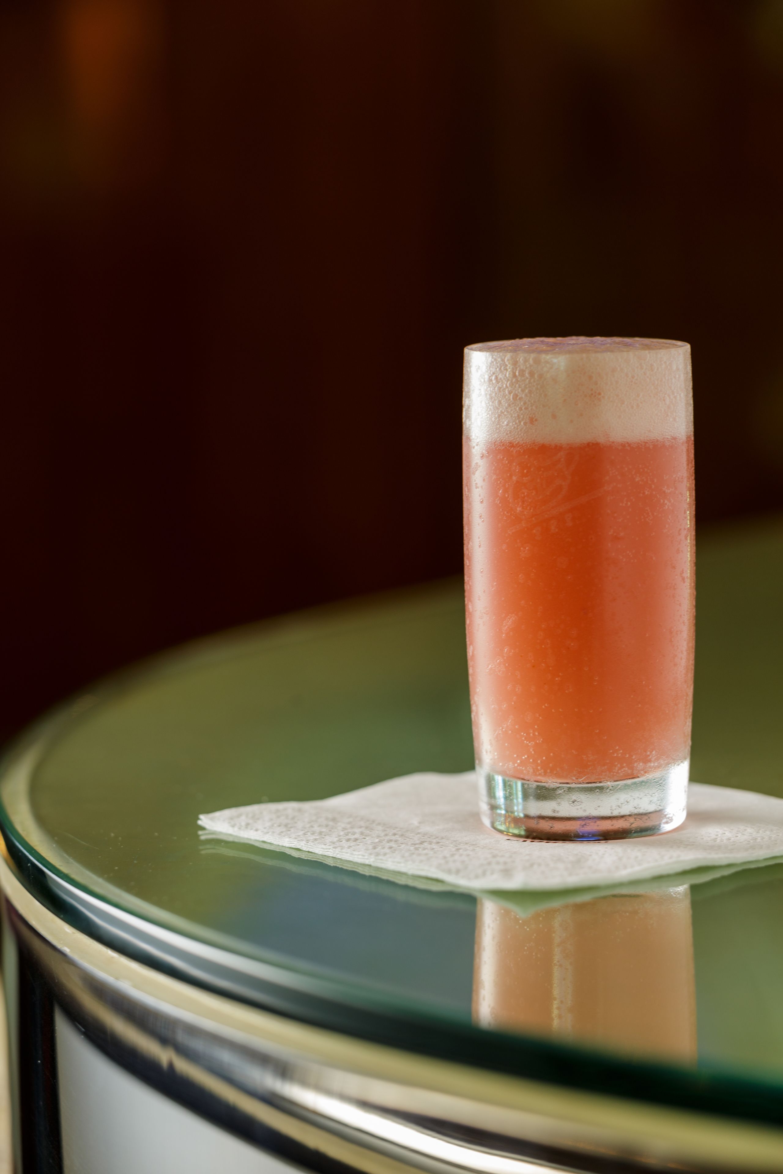 Ein hohes Glas mit einem rosa Getränk auf einer Glastisch mit einer Servietten.