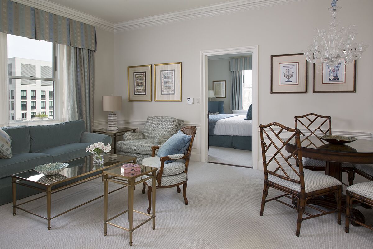 A well-lit living room with a sofa, armchair, coffee table, and dining table. There are framed pictures on the wall and a chandelier.