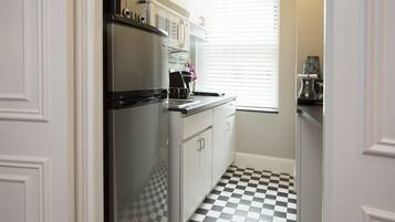 A narrow kitchen with a stainless steel refrigerator, white cabinets, a checkered floor, and a window with blinds.