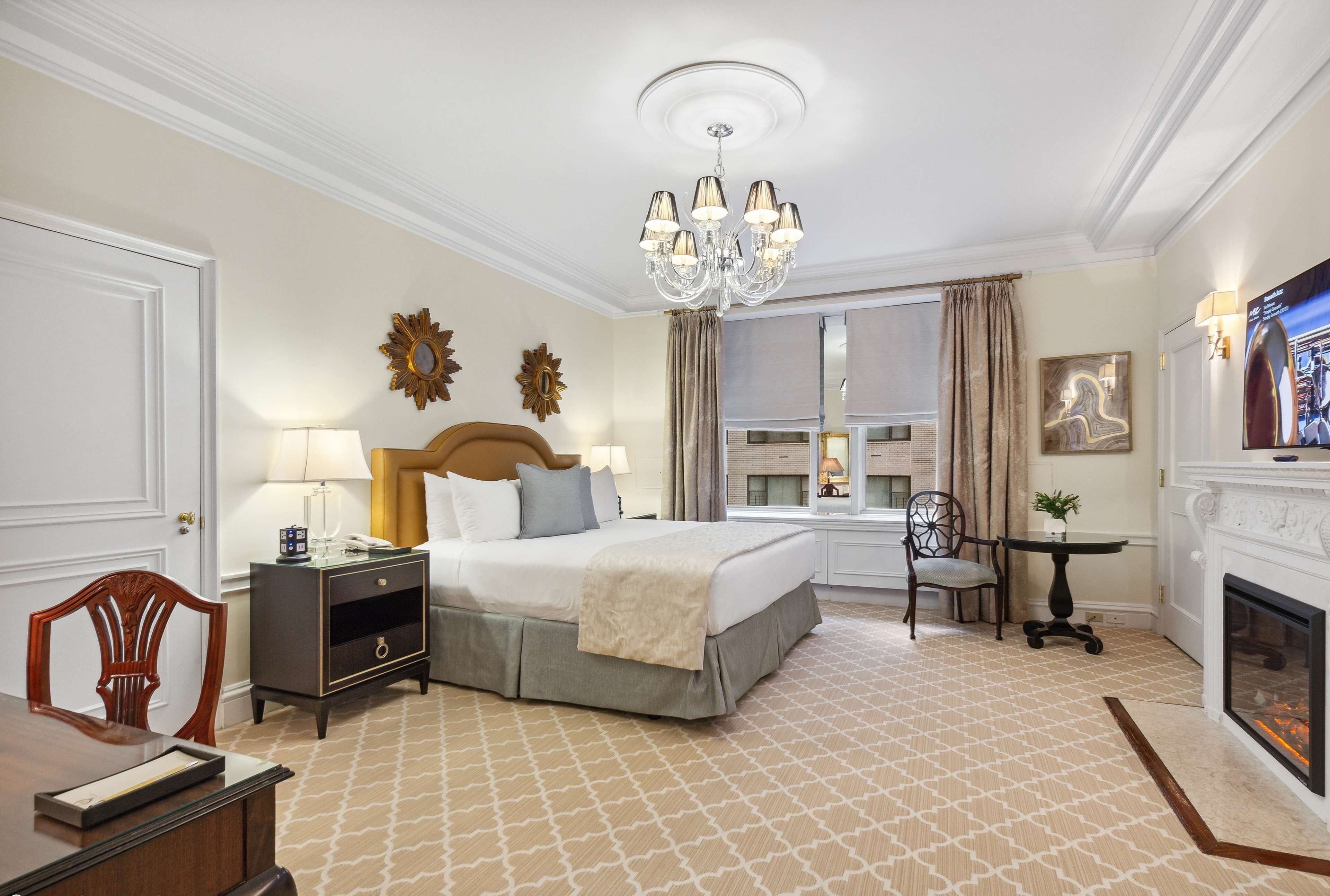 A hotel room with a bed, bedside table, lamp, chair, fireplace, and a window with curtains.