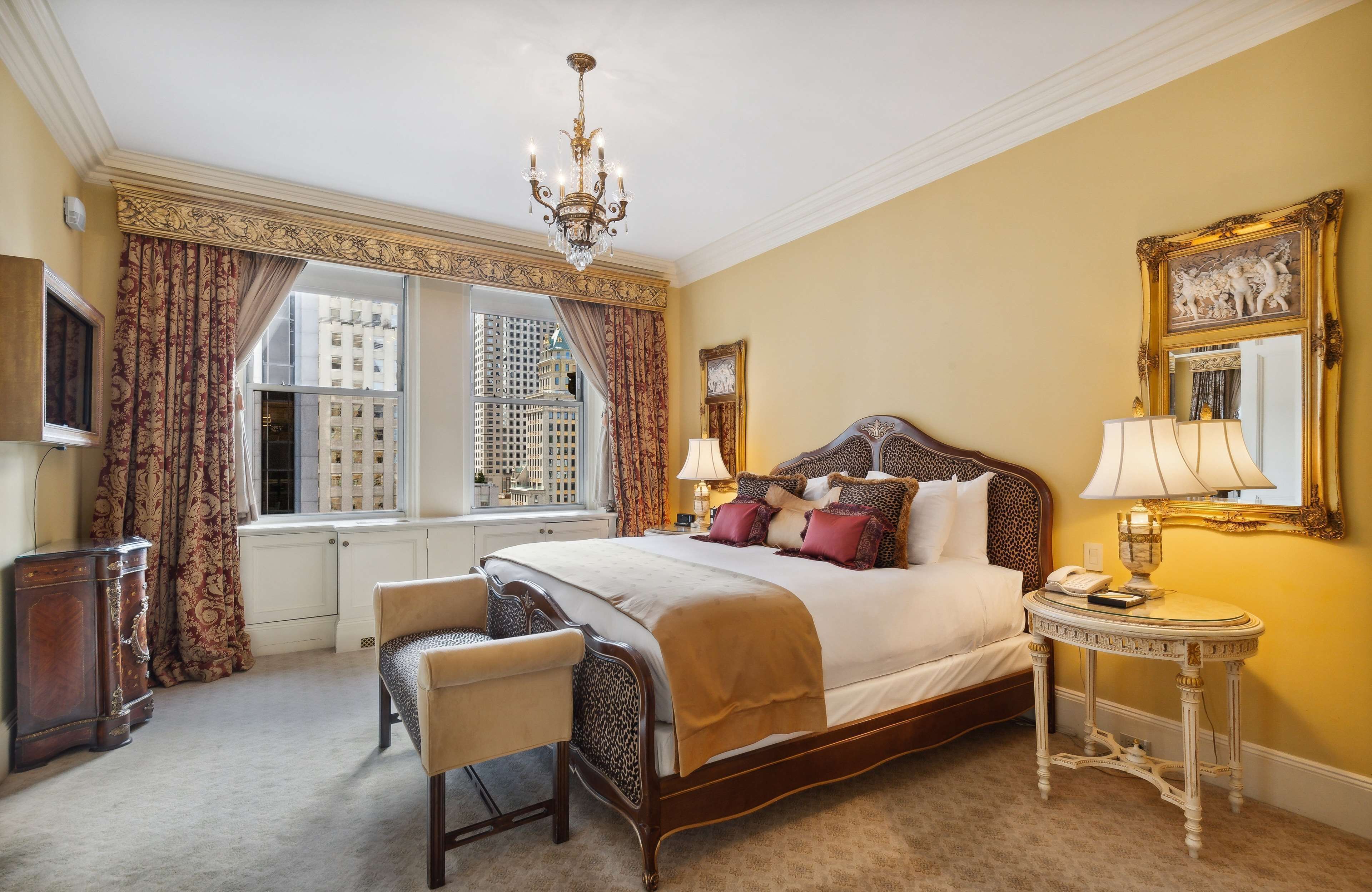 A bedroom with a large bed, a chair, a bedside table with a lamp, a mirror, and a window with curtains.