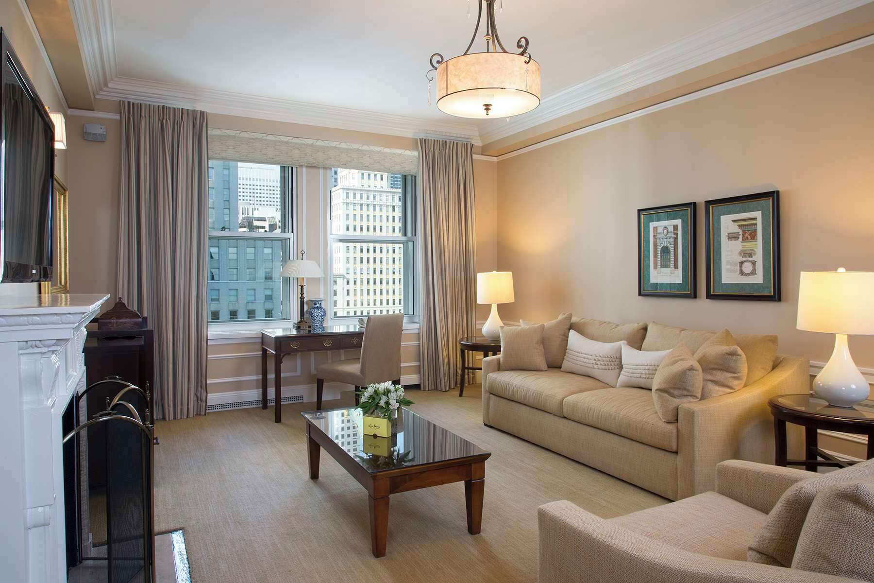 A well-lit living room with a sofa, armchairs, a coffee table, and a fireplace. Large windows offer a view of the cityscape.