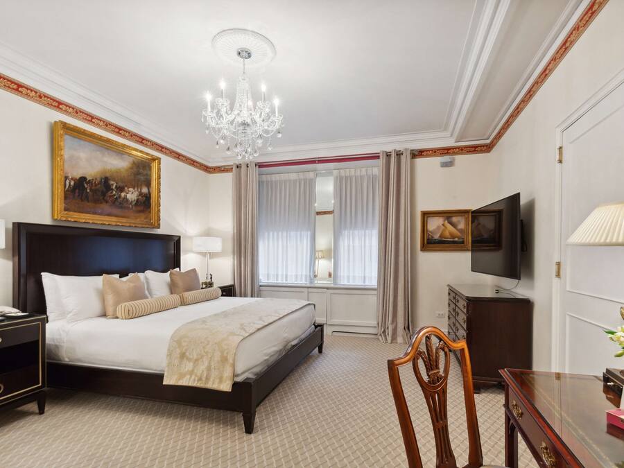 A hotel room with a large bed, a chandelier, a television, a desk with a lamp, and a painting on the wall.