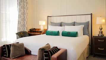 Quarto de hotel com uma cama grande, uma mesa de cabeceira de madeira, uma escrivaninha com abajur e uma poltrona de couro.