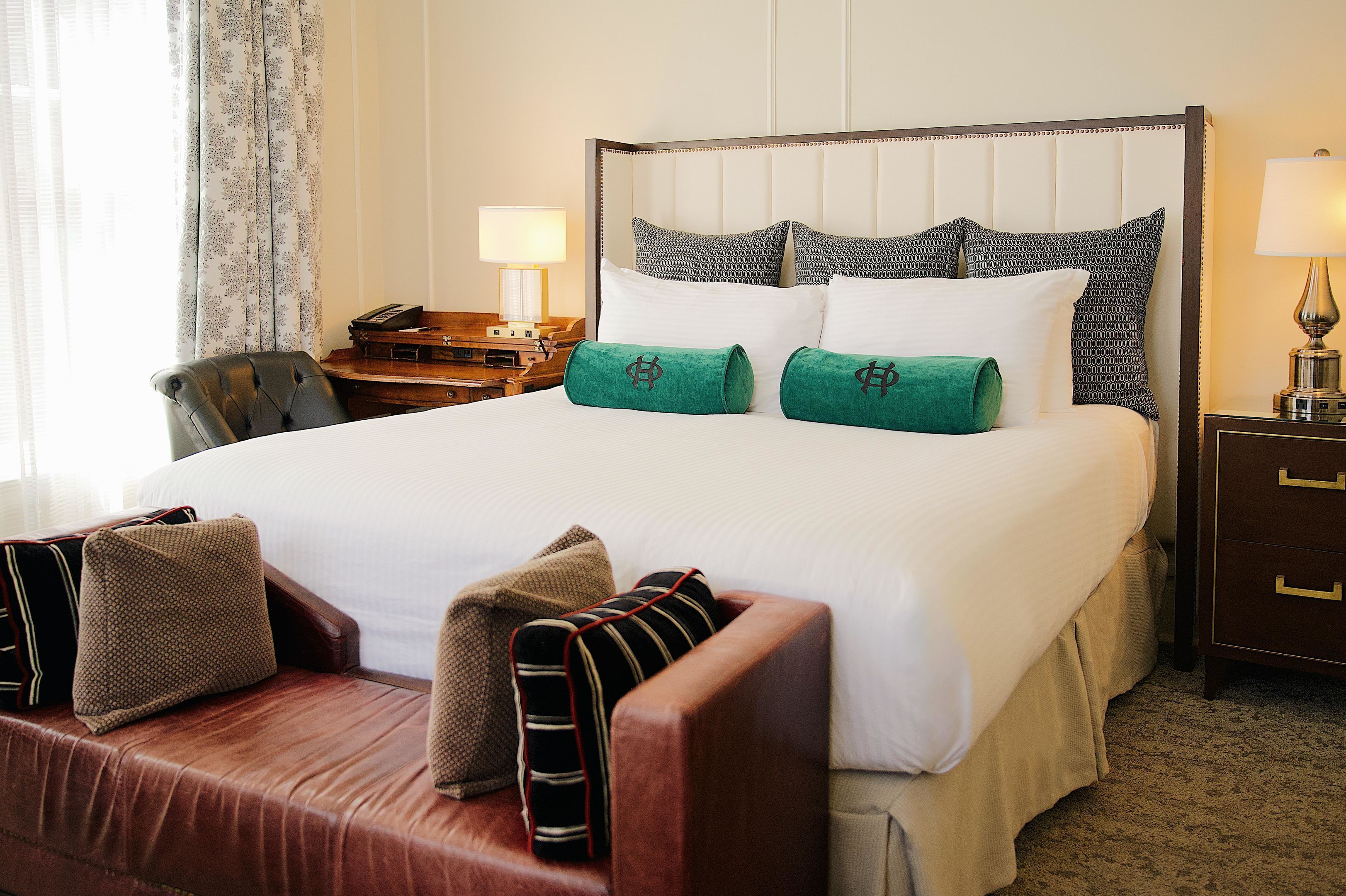 Quarto de hotel com uma cama grande, uma mesa de cabeceira de madeira, uma escrivaninha, uma poltrona de couro e uma janela com cortinas.
