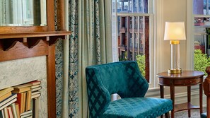 A room with a teal upholstered chair, a wooden side table with a lamp, and a view of buildings through a window. - The Oxford Hotel (Denver)