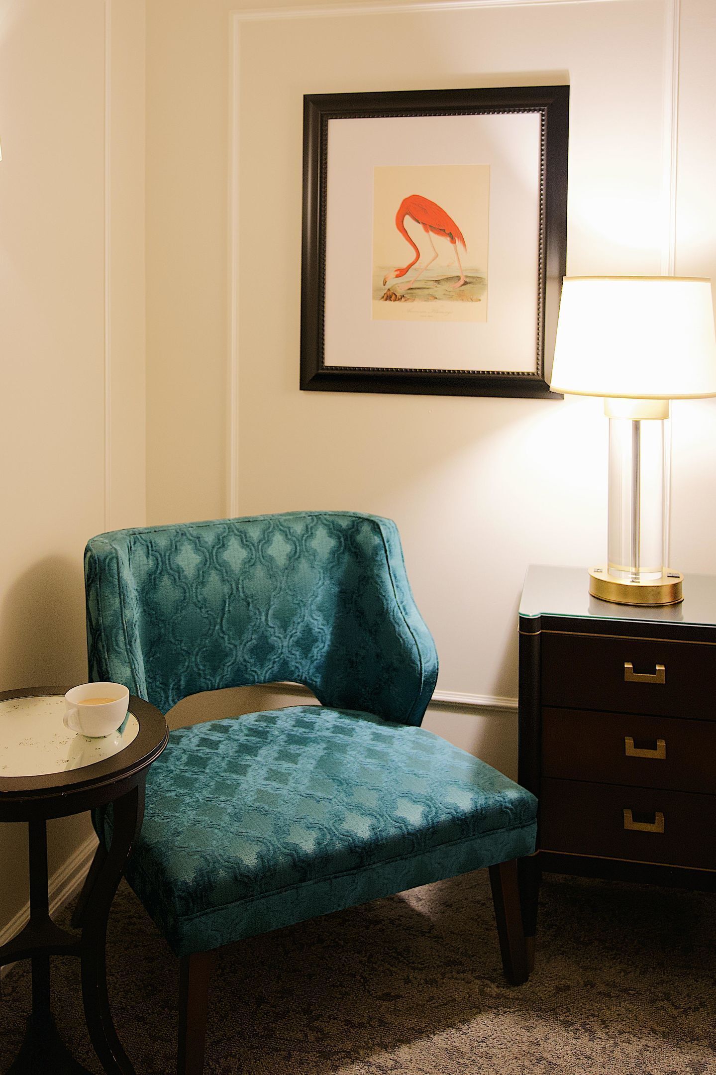 A teal upholstered chair, a small round table with a cup, a framed artwork of a flamingo, and a bedside lamp on a dresser.