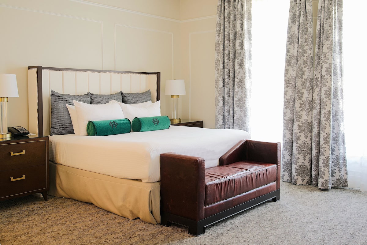 A hotel room with a bed, a nightstand, a lamp, a sofa, and a window with curtains.