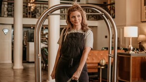 A woman in a hotel lobby with a luggage cart, a chandelier, and a dining area in the background. - The Oxford Hotel (Denver)